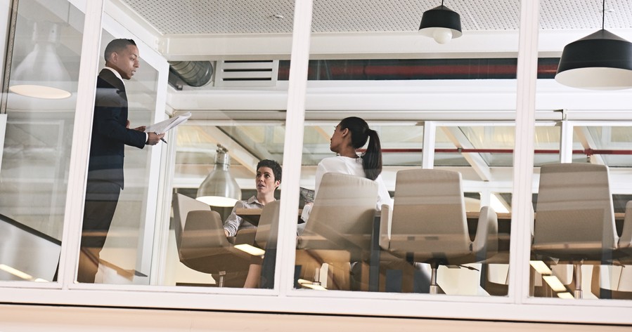 Image is of three people sitting in a modern office near the windows of a conference room.