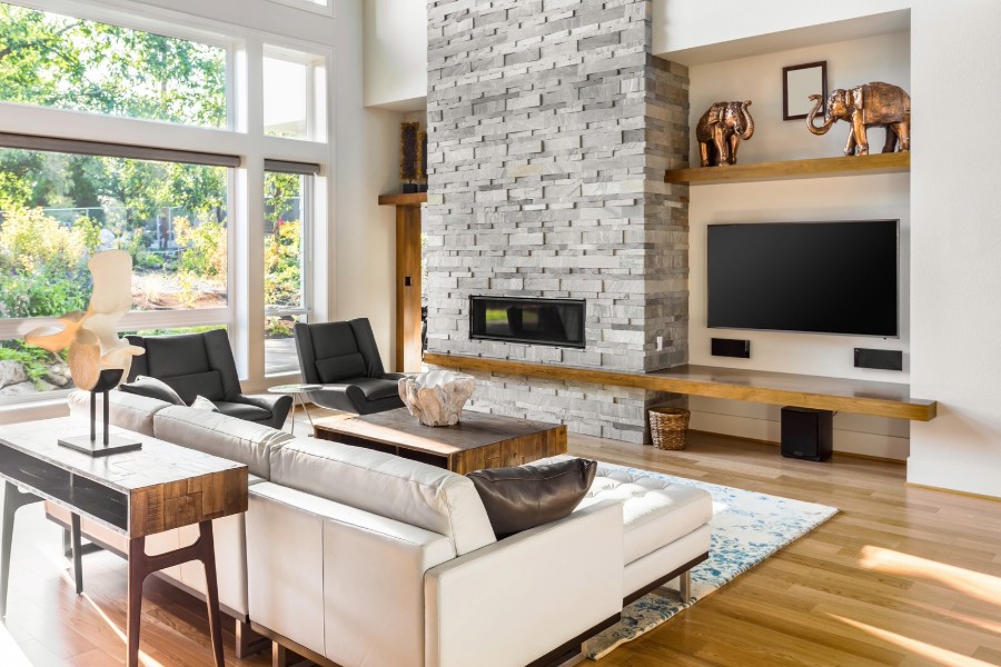 A modern living room with a white couch, wooden side table, stone fireplace, flat-screen TV, and large windows.