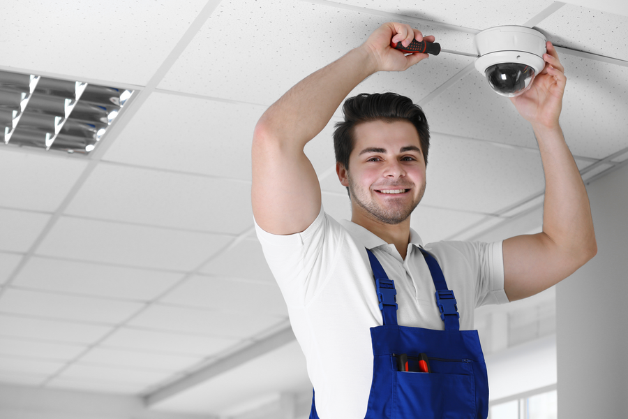 A man in overalls installs a security camera on the ceiling. 