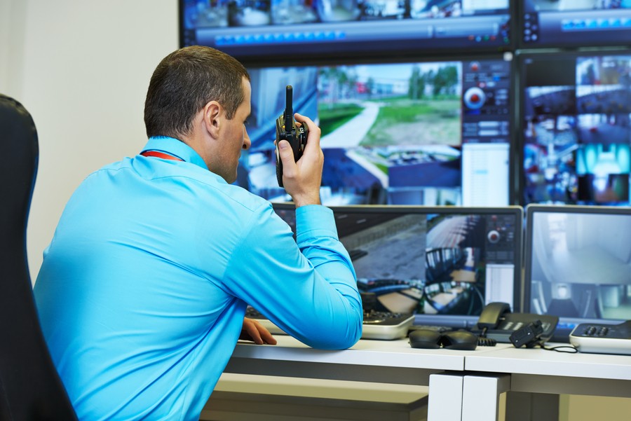 A security professional in a blue shirt uses a walkie-talkie while monitoring multiple surveillance camera feeds on large screens in a control room.