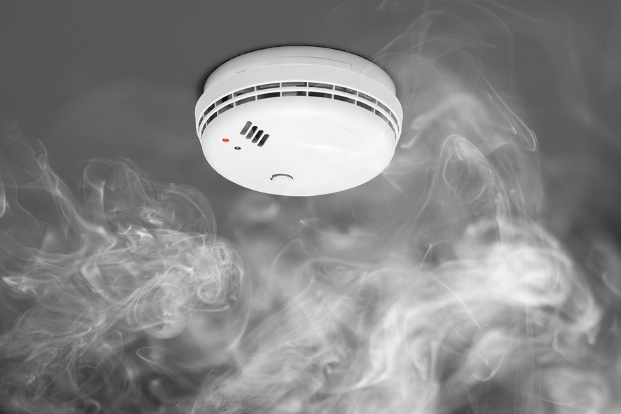 A white smoke detector is mounted on a gray ceiling with visible smoke swirling around it.