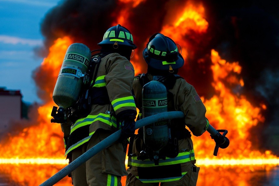 Two firefighters in full gear and breathing tanks holding a hose are viewed from behind with a large fire in front of them. 